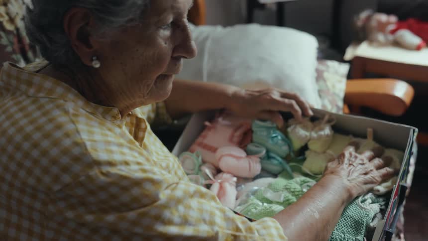 An elderly woman in a yellow checkered shirt sits indoors and looks through a box of baby clothes she gently touches and reorganizes various small colorful knitted items like booties and sweaters
