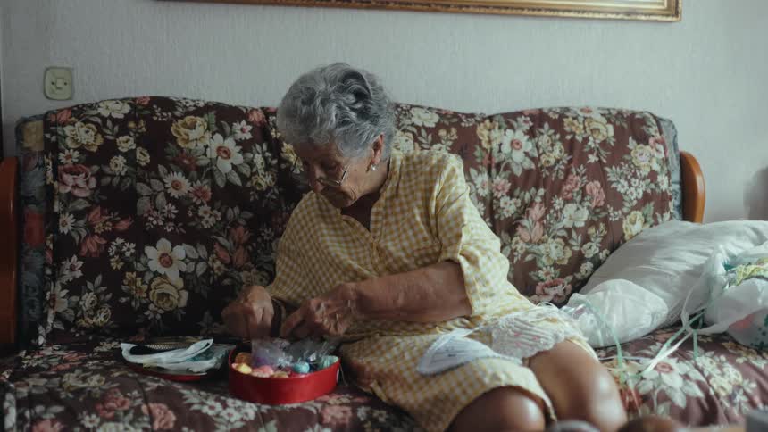 An elderly woman sits on a floral sofa and sorts through a tin of sewing supplies