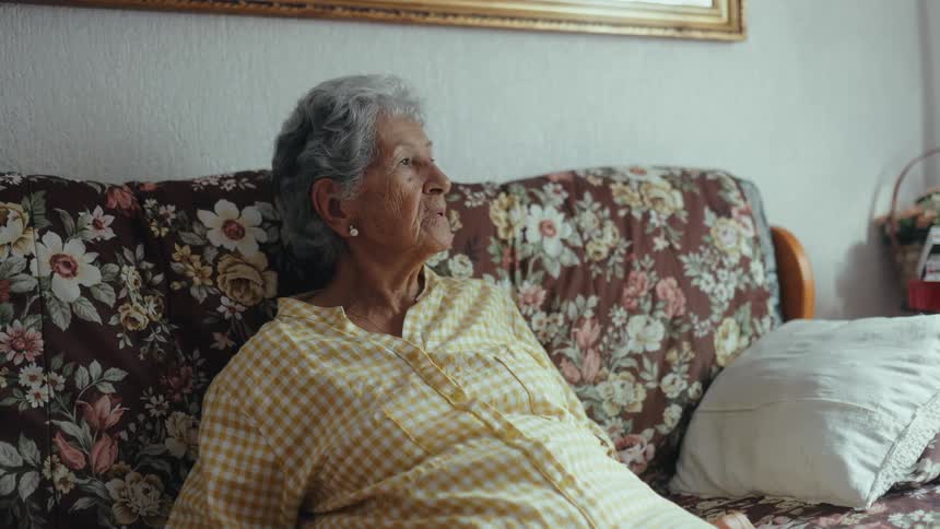 An elderly woman with grey hair and a yellow checkered shirt sits on a floral couch she is looking off to the side with a quiet contemplative expression