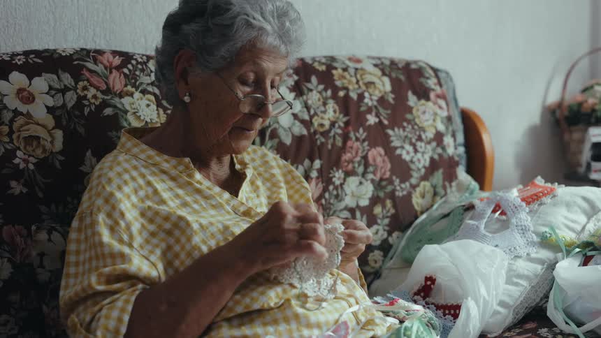 An elderly woman sits on a floral sofa while quietly crocheting a delicate piece of white lace