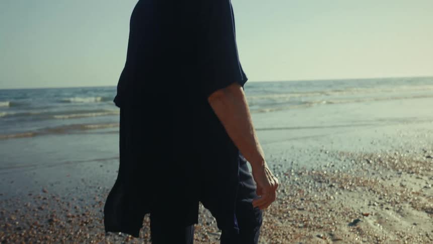 An elderly woman walks along a pebbled beach
