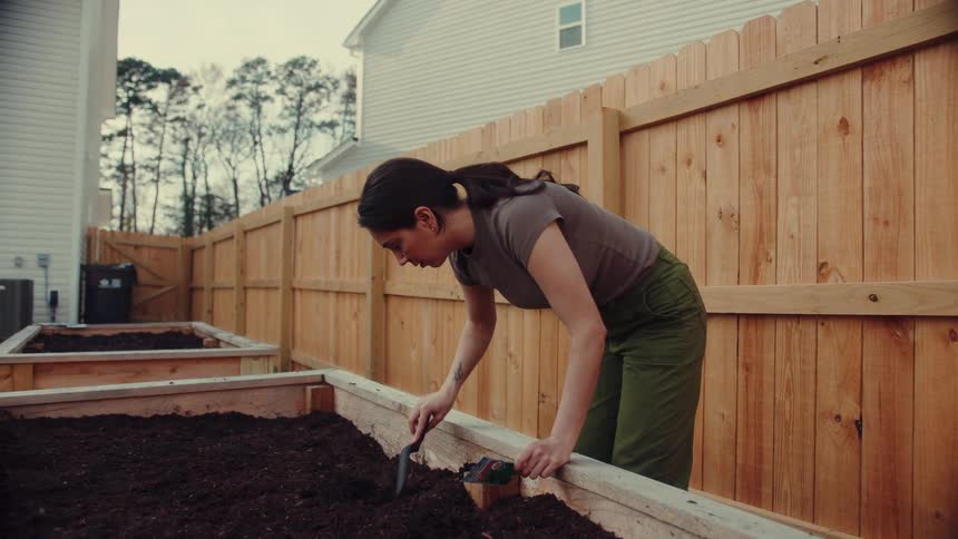 Woman working in a raised soil garden bed in a backyard enclosed by a wooden fence