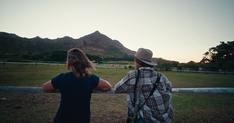 Two people lean against a rail looking at a pasture located near a mountain