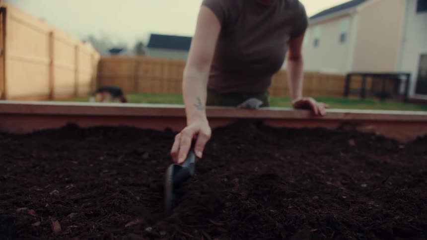 A woman plants seeds in a raised garden bed in her backyard