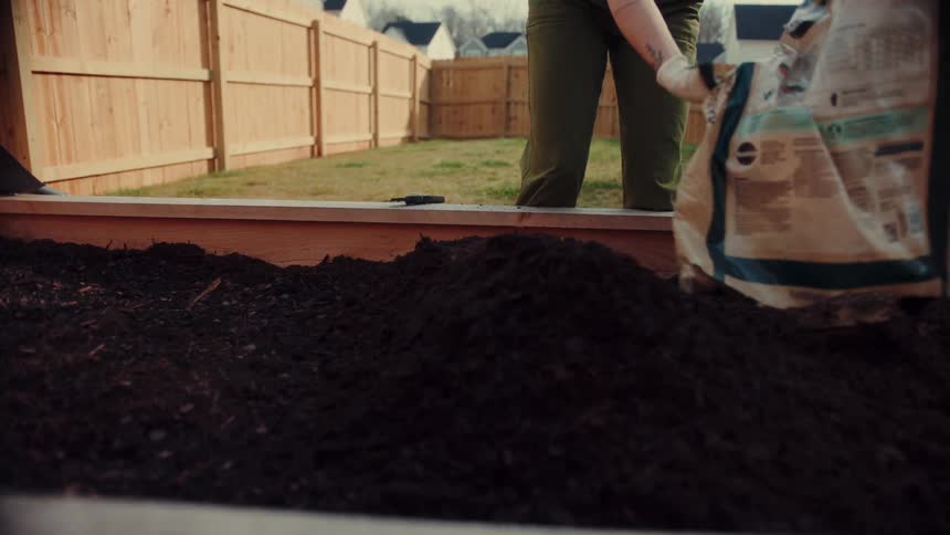 A person wearing gardening gloves pours a large bag of organic raised bed soil into a wooden planter box the scene takes place in a residential backyard ending with the person setting the empty bag aside after filling the bed