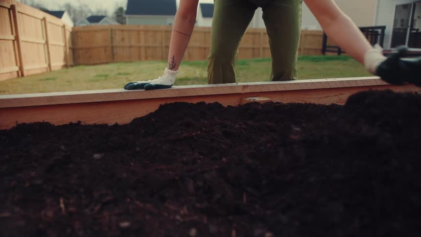 A person wearing protective gloves is shown leveling out dark rich soil within a newly constructed wooden raised garden bedthe scene takes place in a residential backyard with the individual focused on smoothing the earth to prepare it for planting