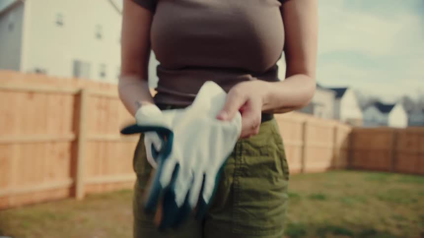 A person stands in a backyard and pulls a pair of white and blue gardening gloves onto their hands the background features a wooden fence and suburban houses under a clear bright sky