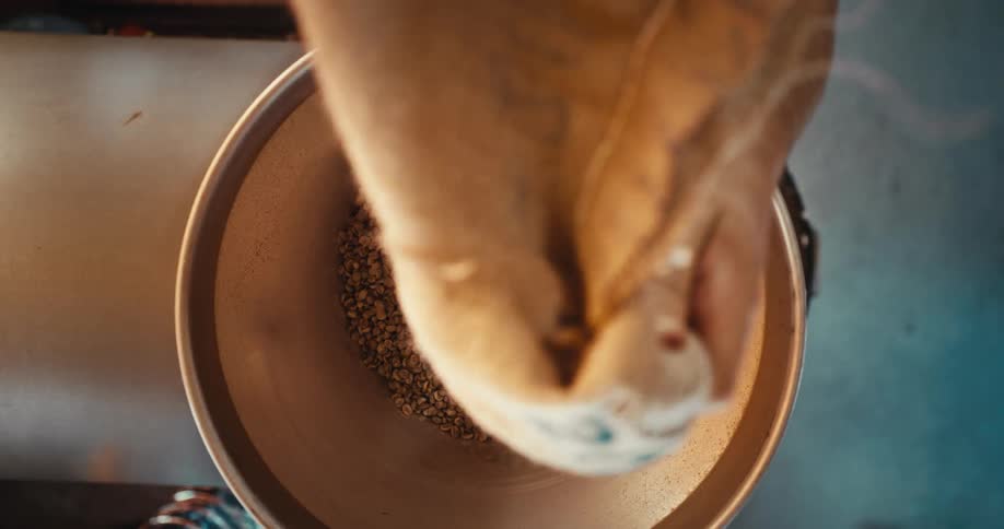 Hands pour a large sack of green unroasted coffee beans into a metal industrial hopper the beans quickly fill the circular container from a topdown perspective until the bag is emptied