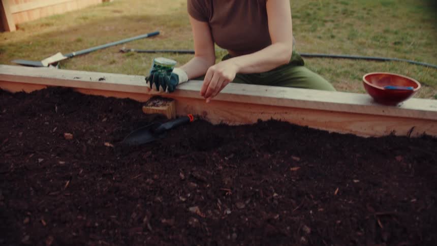 A woman kneels beside a wooden raised garden bed using a small hand trowel to dig a hole in the dark soil she holds a packet of seeds in one hand while carefully placing seeds into the earth and smoothing the dirt over them