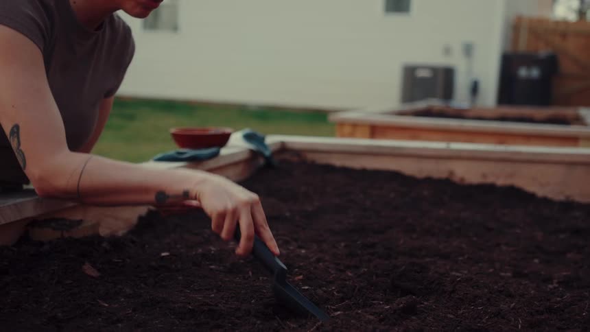 A woman plants seeds in a raised garden bed in her backyard