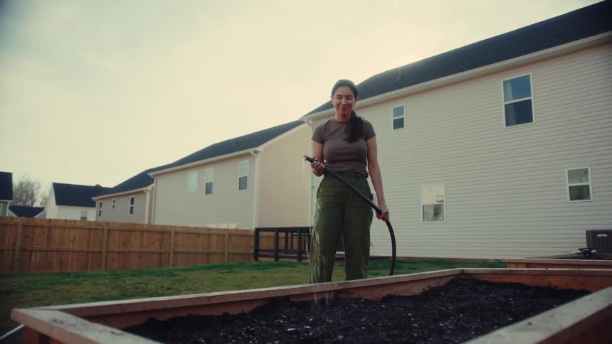 Woman watering a raised soil garden bed with a hose in a residential backyard