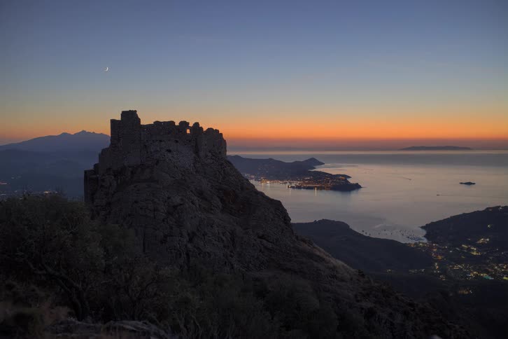 Ancient stone ruins sit atop a rocky cliff overlooking a glowing coastal city and calm sea at dusk
