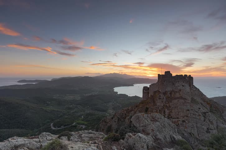 Cars drive through a coastal village beneath a mountaintop castle at sunset