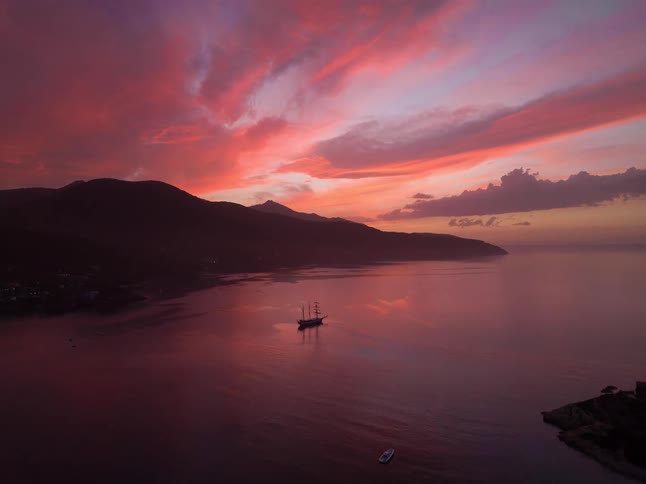 A tall ship floats in the center of a calm bay while a vibrant pink and orange sunset glows over dark mountain silhouettes and a small coastal village as the drone pulls back to reveal the wider harbor at dusk