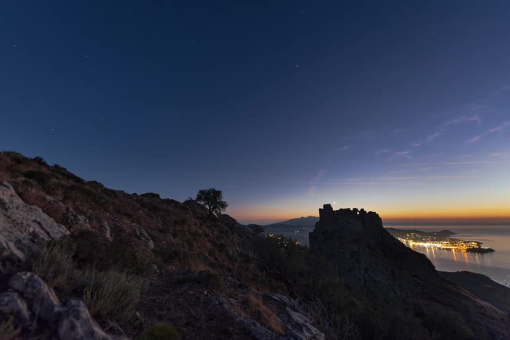A time lapse shows the sun setting over the mediterranean sea and the sant salvador castle ruins in spain as stars appear in the night sky above the glowing lights of a coastal town