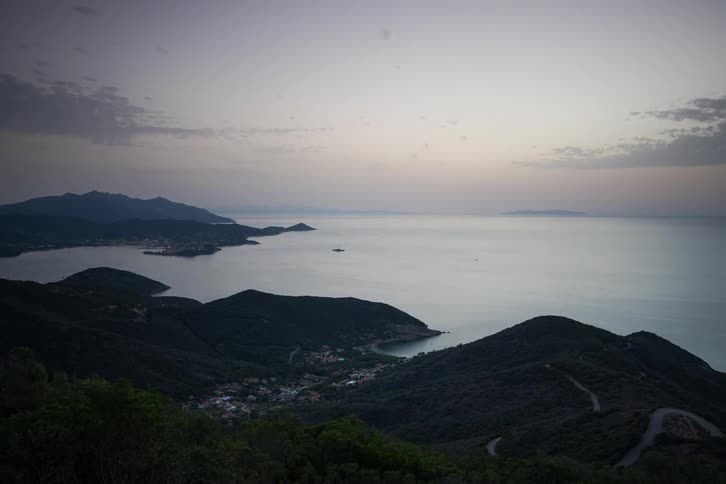 A time lapse view shows a coastal bay with rolling hills as the sun sets over the ocean while light clouds drift across the sky and small towns begin to glow with golden lights during the twilight