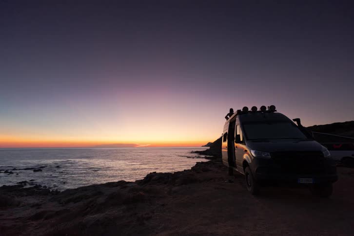 A black van sits on a dirt cliff by the ocean during a timelapse that transitions from a golden sunset into a starry night sky as the moon rises over the water and people move around the vehicle