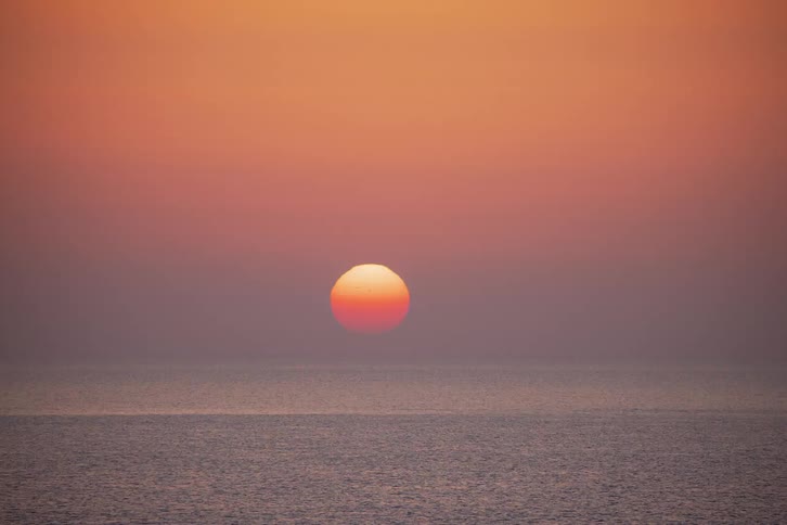 A time lapse shows a large orange sun slowly sinking below the horizon of a calm sea under a clear gradient sky during a warm sunset