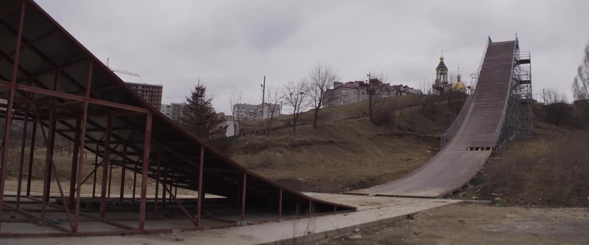 A large weathered wooden ski jump ramp sits on a grassy hillside under a cloudy skyin the background a golddomed church and several residential buildings are visible across the landscape