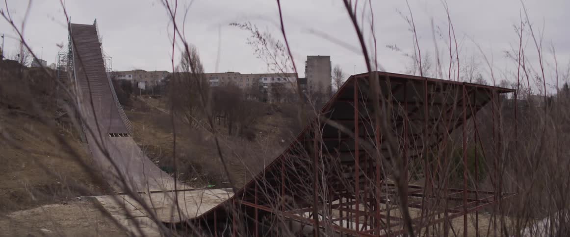 An abandoned and overgrown ski jump ramp stands derelict in a desolate urban landscape under an overcast sky