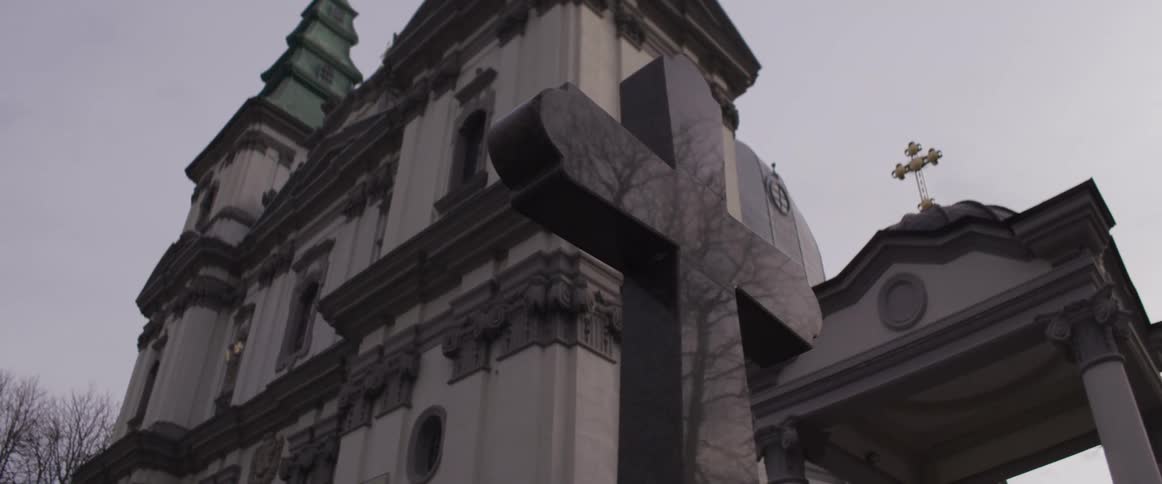 A low view shows a black marble cross in the foreground with the ornate facade and green spires of the cathedral of the immaculate conception of the holy mother of god in ternopil under a grey sky