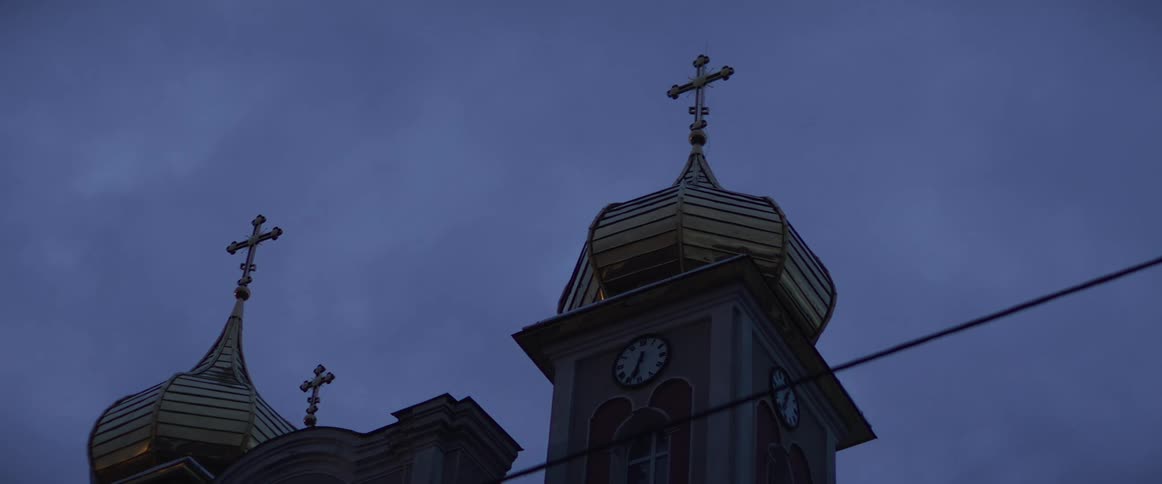 The camera pans upward toward the golden onion domes and ornate crosses of an orthodox church against a dark blue twilight sky capturing the detailed clock face on the side of the bell tower as power lines stretch across the lower foreground