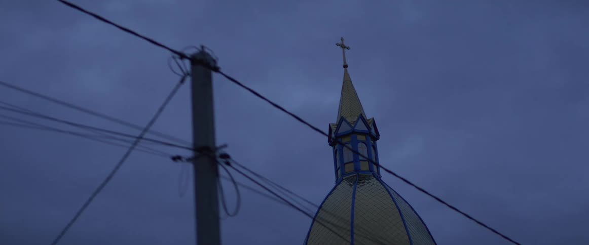 A church steeple and cross are seen through power lines against a dark twilight sky