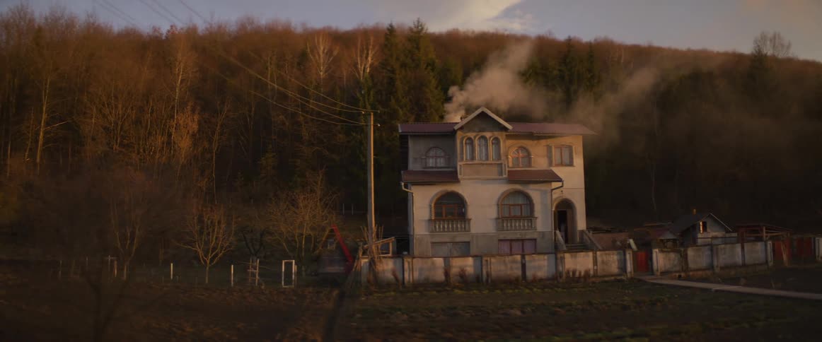 A twostory house with arched windows and a smoking chimney sits at the edge of a dense autumnal forestthe camera captures a passing view of the propertys fenced yard and surrounding fields under the warm glow of late afternoon light