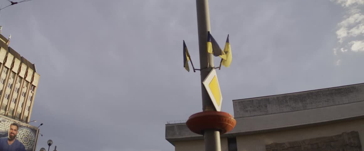 The video shows a street scene looking up at a utility pole against a cloudy sky here are two lines describing the visualsmall ukrainian flags flutter from a metal pole that also features a yellow diamondshaped priority road signas th