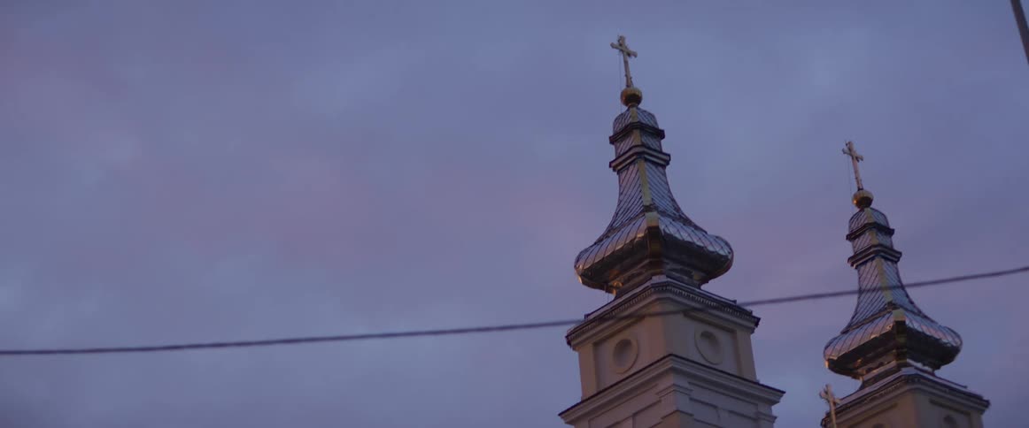 The camera pans past ornate church steeples with shining silvertiled domes and golden crosses against a dusky sky fading daylight illuminates the architecture and nearby power lines while soft pink clouds drift in the background