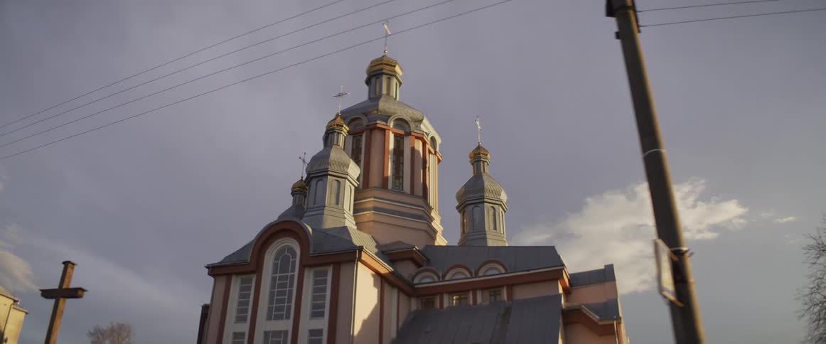 A large orthodox church with multiple silver and gold domes stands against a pale blue sky while power lines and a wooden pole pass through the foreground of the moving shot