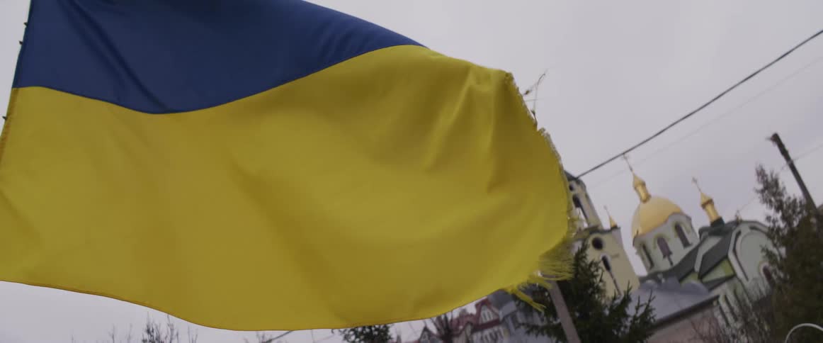 The tattered blue and yellow flag of ukraine ripples in the wind while a traditional church with golden domes stands under a gray overcast sky in the background