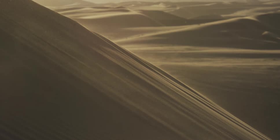 Golden sunlight catches the fine mist of sand blowing off the sharp crest of a desert dune in the background a vast landscape of rolling sand hills stretches toward a hazy warm horizon
