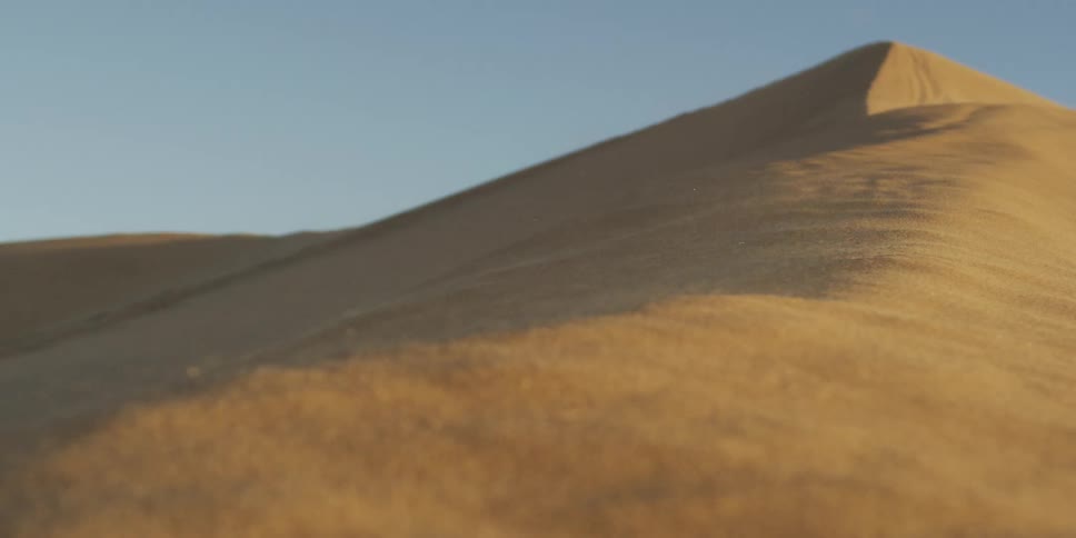 The video shows a closeup groundlevel view of wind blowing sand across the surface of a large desert dune in the background the peak of the dune stands sharply against a clear light blue sky under bright daylight