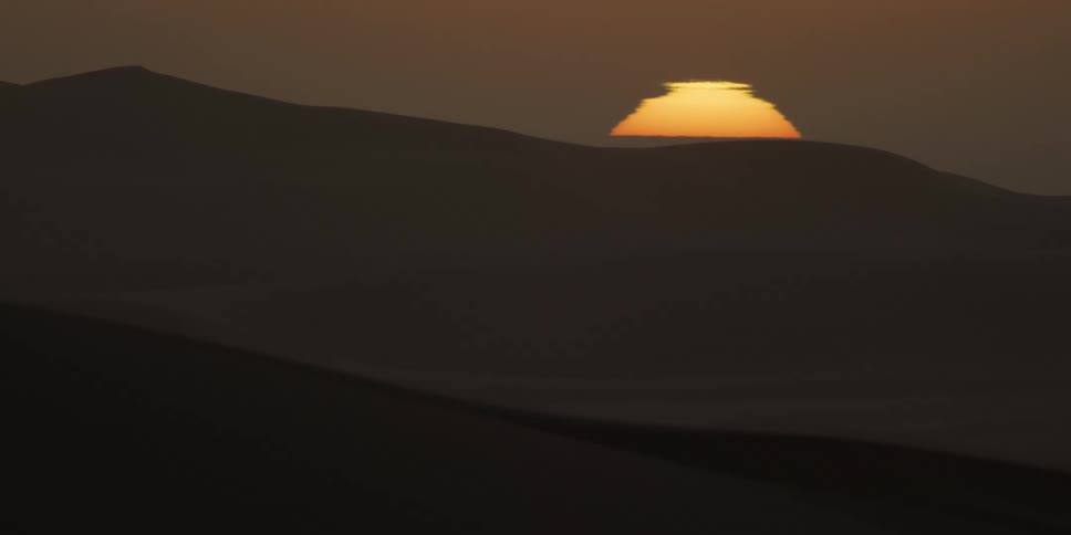 The bright orange sun sets slowly behind the dark silhouettes of rolling sand dunes in the sahara desert under a hazy twilight sky