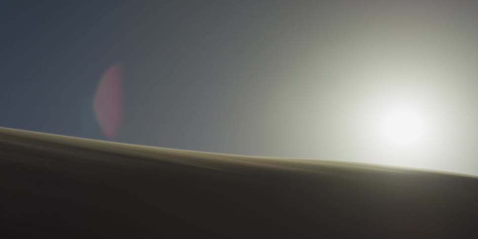A bright sun hangs in a pale sky over the crest of a large sand dune in the sahara desert as fine sand particles across the dark ridge in the foreground