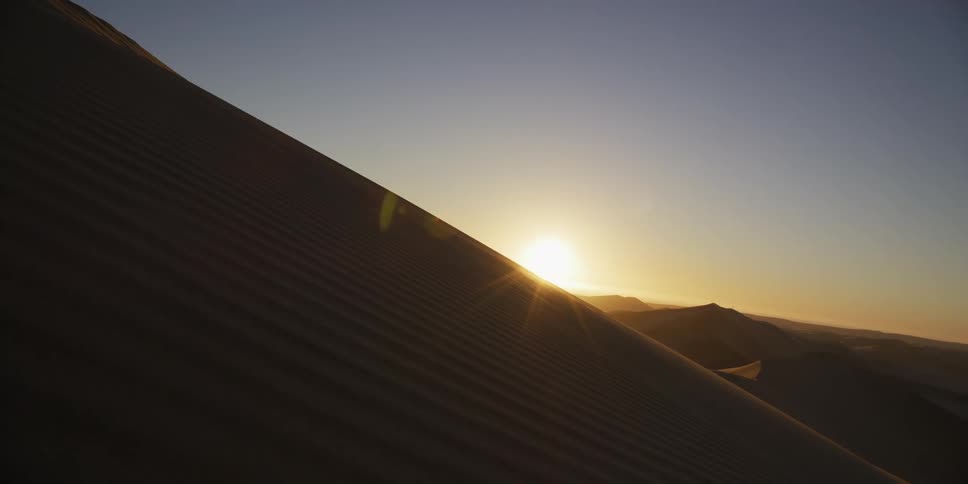 The sun crests over the ridge of a large sand dune in the sahara desert at dawn with golden light rays and lens flare against a clear blue sky