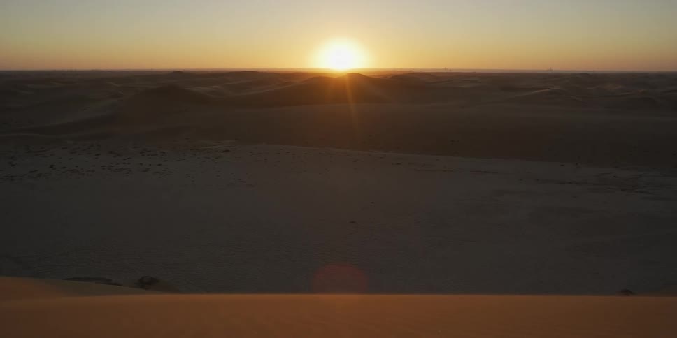 The sun sits low on the horizon casting a brilliant orange glow and long shadows across an endless expanse of desert dunes a soft lens flare catches the light in the foreground highlighting the tranquil and vast nature of the arid landscape