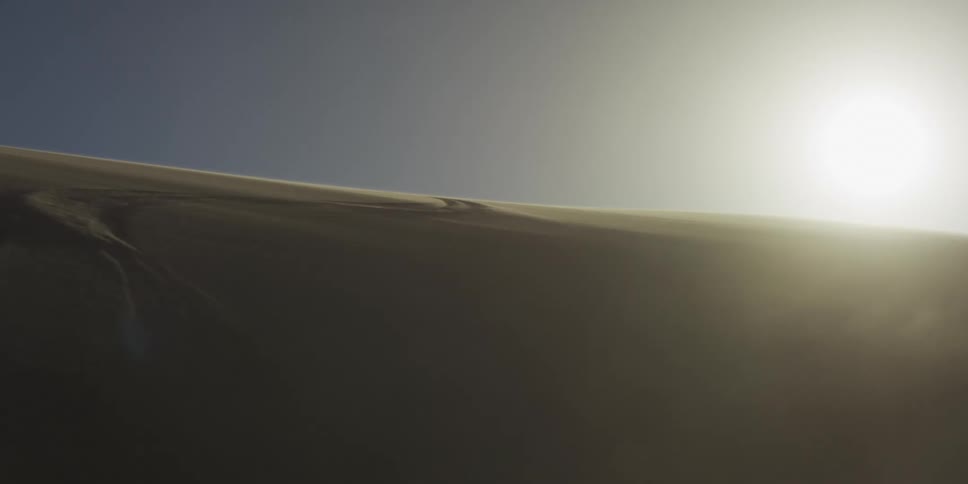 A bright sun shines in a hazy sky over a large sand dune in the sahara desert as wind gusts thick clouds of sand across the dark slopes in the foreground