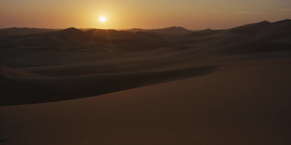 An aerial view shows the sun rising over the vast sand dunes of the sahara desert as golden light spreads across the rolling landscape under a clear sky