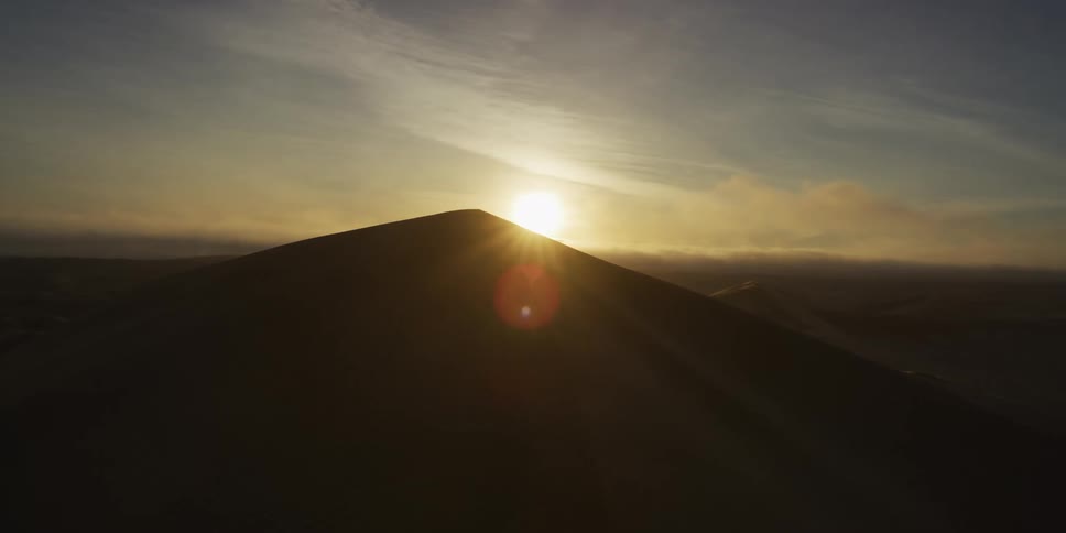 An aerial shot captures the sun rising directly behind the sharp peak of a sand dune casting a warm golden glow over the desert landscapethe light creates a dramatic silhouette of the dune while a faint mist or dust cloud drifts across the horizon in