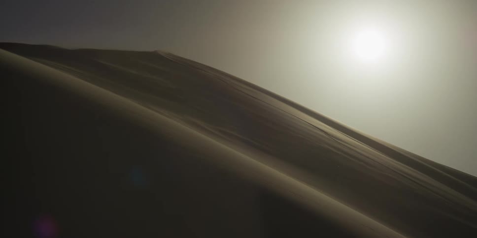 A bright sun shines over a massive sand dune in the sahara desert as wind blows sand across the ridgeline under a hazy and warm sky
