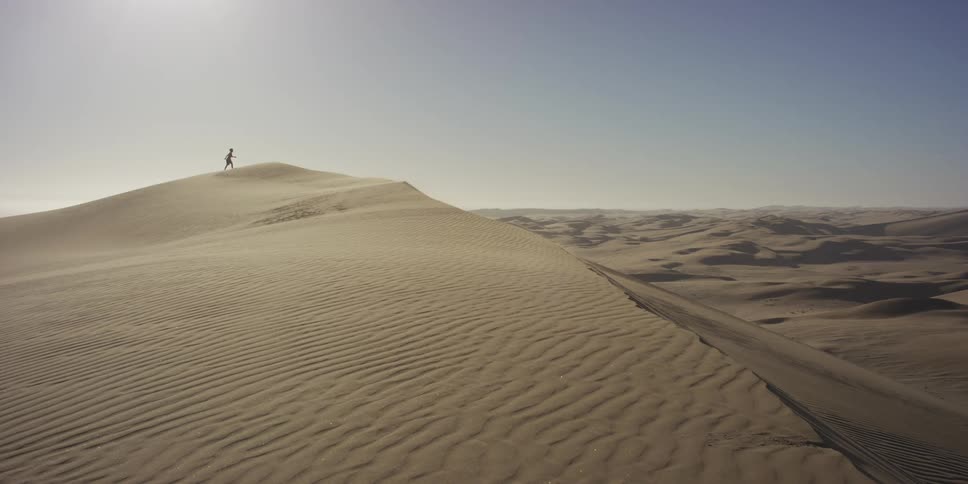 A person climbs a massive sand dune while the sun shines over a vast desert filled with endless rolling ridges and intricate wind patterns on the ground