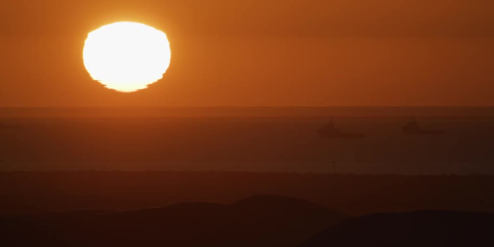 A large bright sun hangs low in a vibrant orange sky over the sea where silhouettes of ships are visible on the horizon beyond the dark outlines of sand dunes