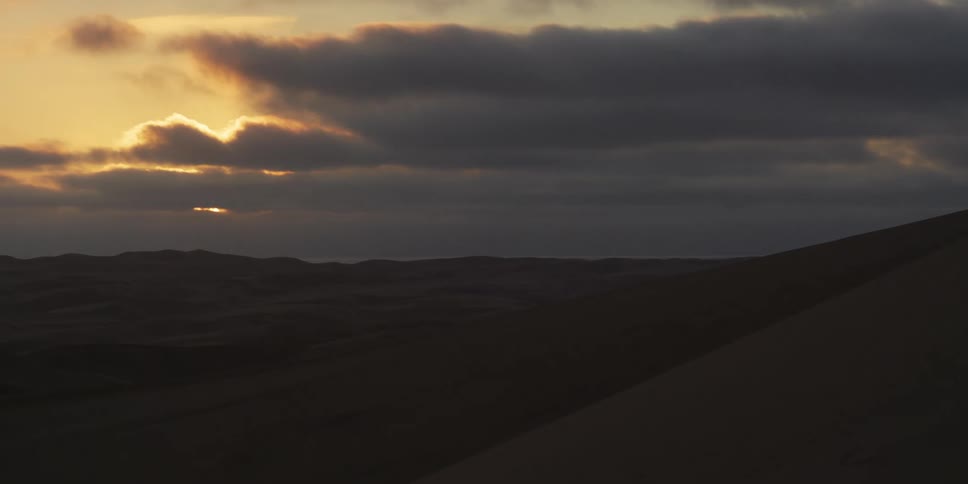 The sun sets behind a layer of thick clouds over a vast desert landscape where dark shadows stretch across the rolling sand dunes and light rays pierce through the hazy sky