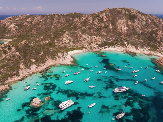 An aerial drone shot captures numerous white boats and yachts anchored in a vibrant turquoise bay surrounded by rugged tan cliffs and a small sandy beach under a clear bright sky