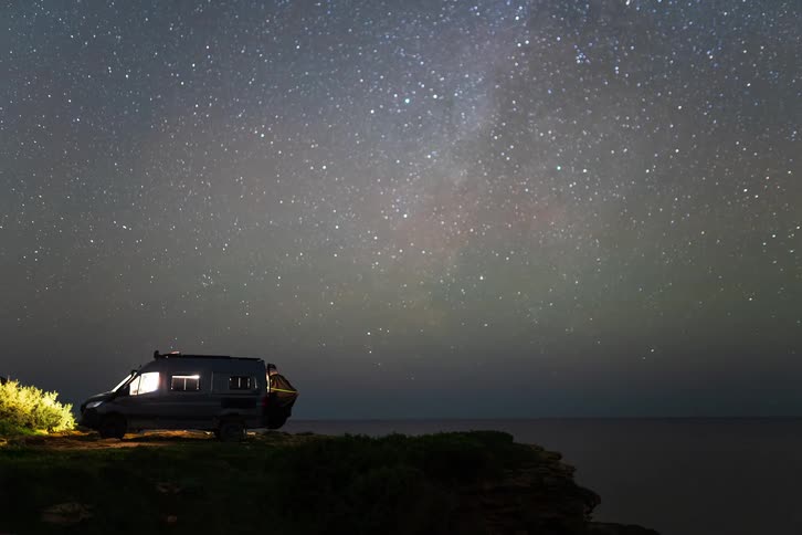 A dark camper van sits parked on a grassy cliff edge at night while the milky way galaxy and thousands of stars stretch across the clear sky above a calm and black ocean
