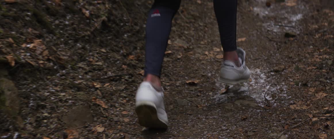 A person wearing dark athletic leggings and white sneakers walks along a narrow leafcovered dirt trail through a wooded area their steps moving steadily forward over the uneven ground dampened by patches of water