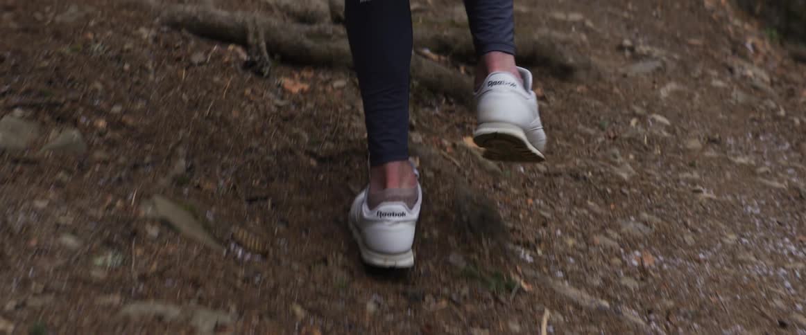 A woman wearing white sneakers and black athletic leggings runs up a steep dirt trail filled with thick tree roots and dry forest debris