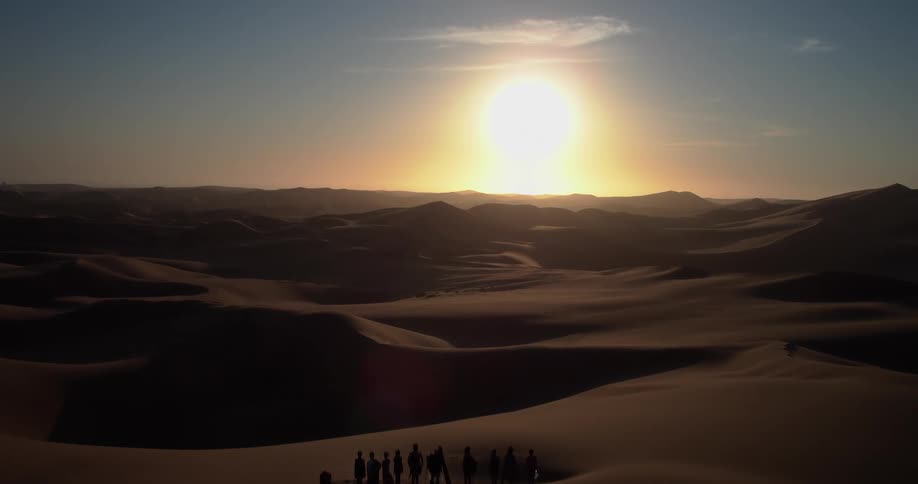 Sun sitting near the red horizon shines across the desert casting people standing on a dune into silhouettes